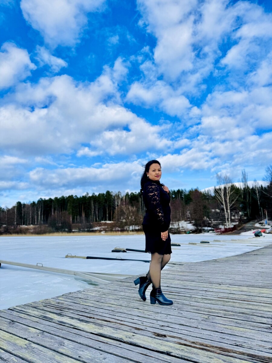 Mustaan mekkoon ja saappaisiin pukeutunut nainen seisoo puisella laiturilla jäätyneen järven rannalla, ja taustalla on metsä ja sininen taivas, jossa on hajapilviä.