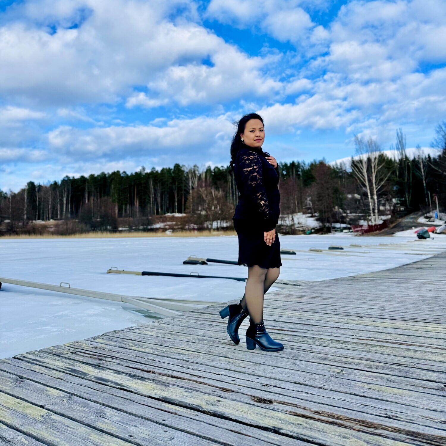 Mustaan mekkoon ja saappaisiin pukeutunut nainen seisoo puisella laiturilla jäätyneen järven rannalla, ja taustalla on metsä ja sininen taivas, jossa on hajapilviä.