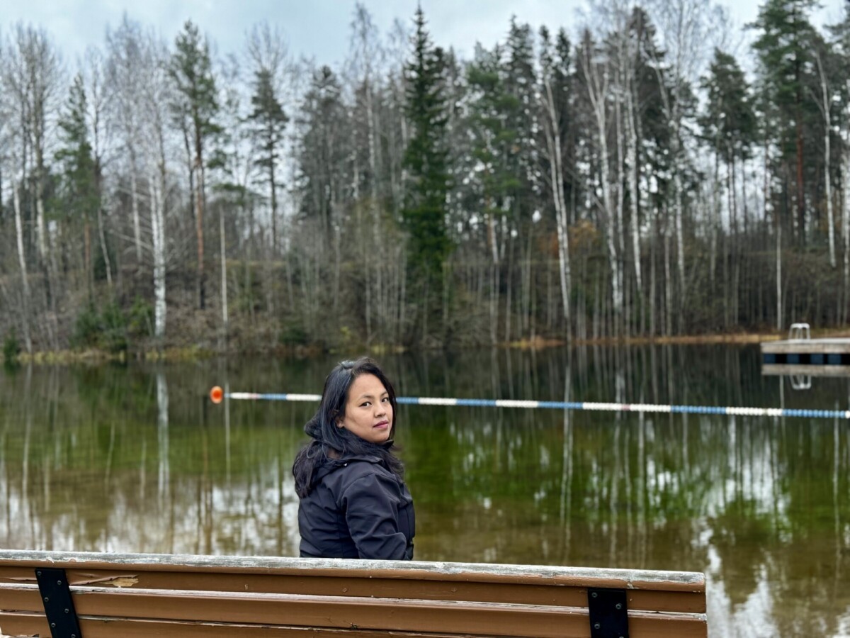 Mustaan takkiin pukeutunut nainen istuu puisella penkillä rauhallisen järven rannalla ja katsoo olkansa yli. Hänen takanaan syksyiset puut ja kelluva pato ulottuvat veden yli pilvisen taivaan alla.