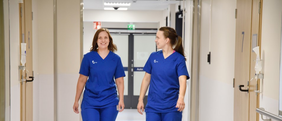 Kaksi sinisiin hoitohaalareihin pukeutunutta naista kävelee sairaalan valaistua käytävää pitkin hymyillen toisilleen. Käytävällä on useita ovia ja kyltti ulospääsyn yläpuolella.