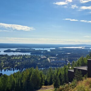 Näkymä kukkulan rinteeltä, josta avautuu näkymä metsään, useisiin järviin, hajallaan oleviin rakennuksiin ja kaukana oleviin mataliin kukkuloihin kirkkaan sinisellä taivaalla, jolla on joitakin pilviä.
