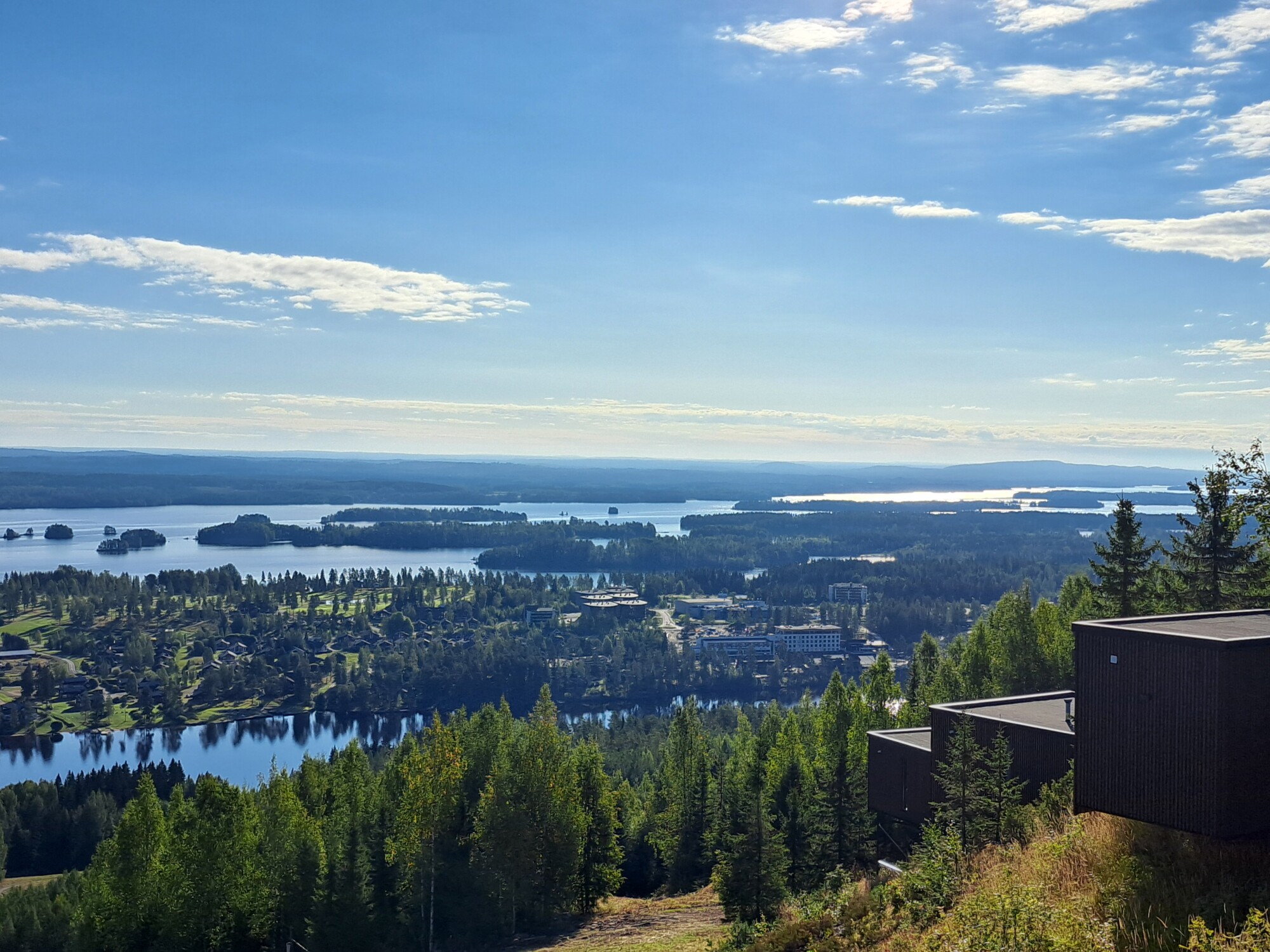 Näkymä kukkulan rinteeltä, josta avautuu näkymä metsään, useisiin järviin, hajallaan oleviin rakennuksiin ja kaukana oleviin mataliin kukkuloihin kirkkaan sinisellä taivaalla, jolla on joitakin pilviä.