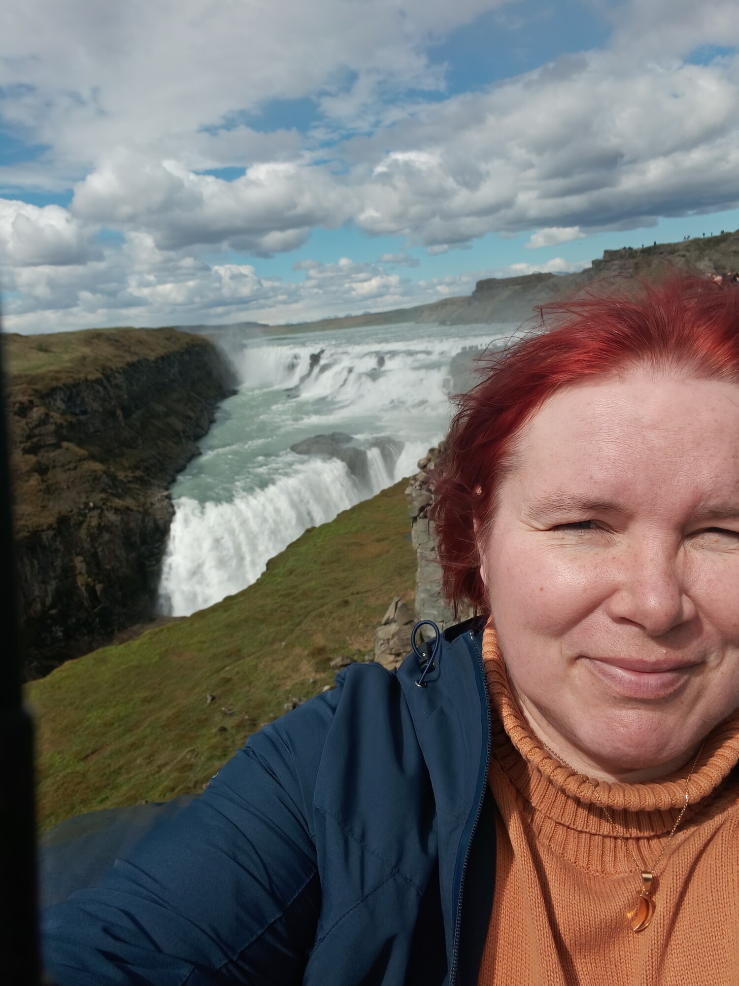 Punatukkainen henkilö, jolla on sininen takki ja oranssi villapaita, ottaa selfien suuren, voimakkaan vesiputouksen edessä, jota ympäröivät vihreät kalliot osittain pilvisen taivaan alla.