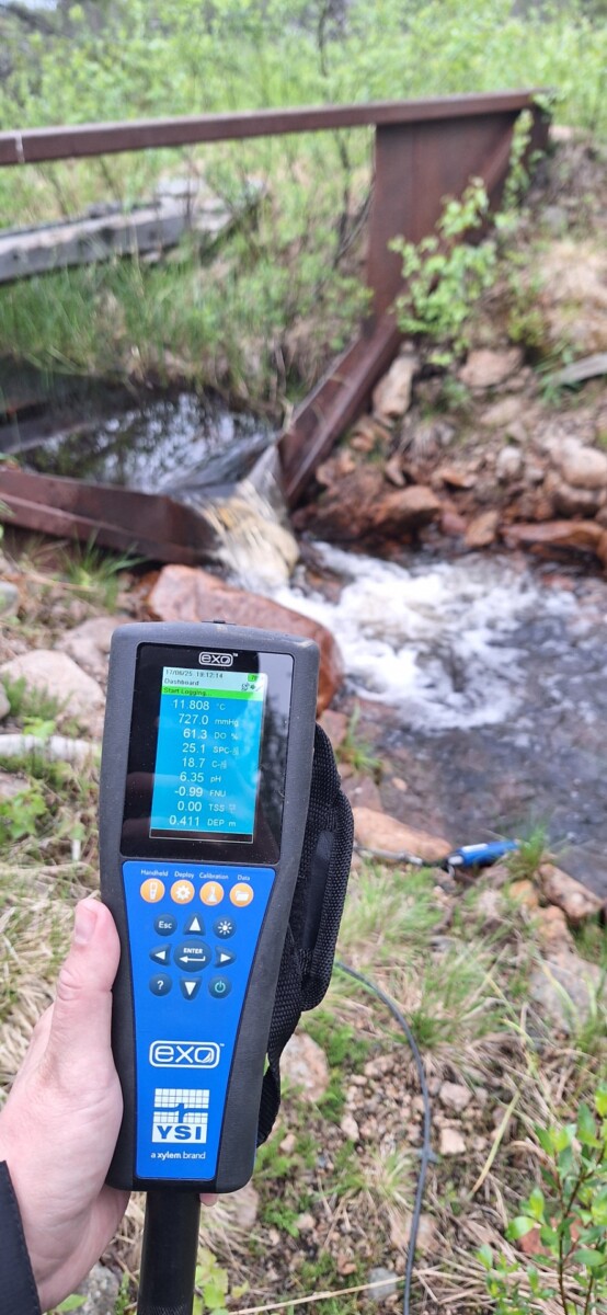 Henkilö pitää kädessä kannettavaa vedenlaatumittaria lähellä pientä puroa, joka virtaa ruosteisen metallisillan alla vihreässä, luonnollisessa ulkoilmaympäristössä. Laite näyttää mittaustiedot näytöllään.