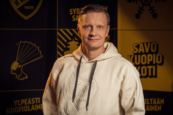 Urheilulogolla varustettu vaaleaan huppariin pukeutunut mies seisoo keltaisen ja mustan taustan edessä, jossa on suomenkielistä tekstiä ja grafiikkaa. Hän hymyilee ja katsoo kameraan.