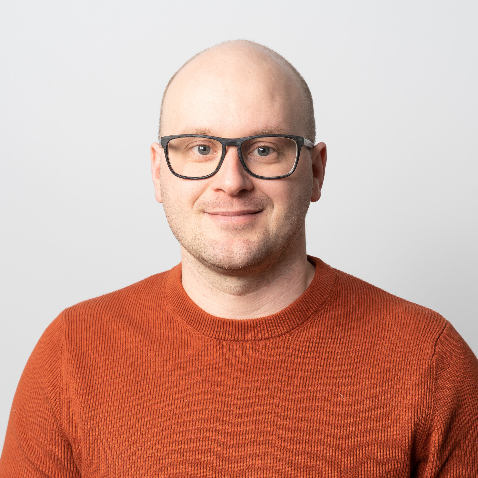 Mies, jolla on ajeltu pää, mustat silmälasit ja oranssi kuvioitu pusero, hymyilee pehmeästi katsoen kameraan vaaleanharmaalla taustalla.