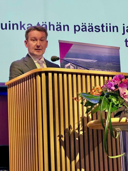 Pukumies puhuu kukkien koristelemalla puisella korokkeella. Hänen takanaan on näyttö, jossa näkyy suomenkielistä tekstiä ja maisemakuvalla varustettu banneri.