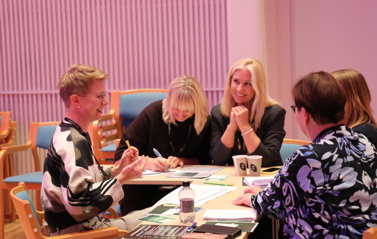 Viisi naista istuu pöydän ääressä ja keskustelee vilkkaasti, hymyilee ja kirjoittaa muistiinpanoja. Pöydällä on papereita, esitteitä, kahvikuppeja ja vesipullo. Ilmapiiri vaikuttaa lämpimältä ja yhteistyökykyiseltä.