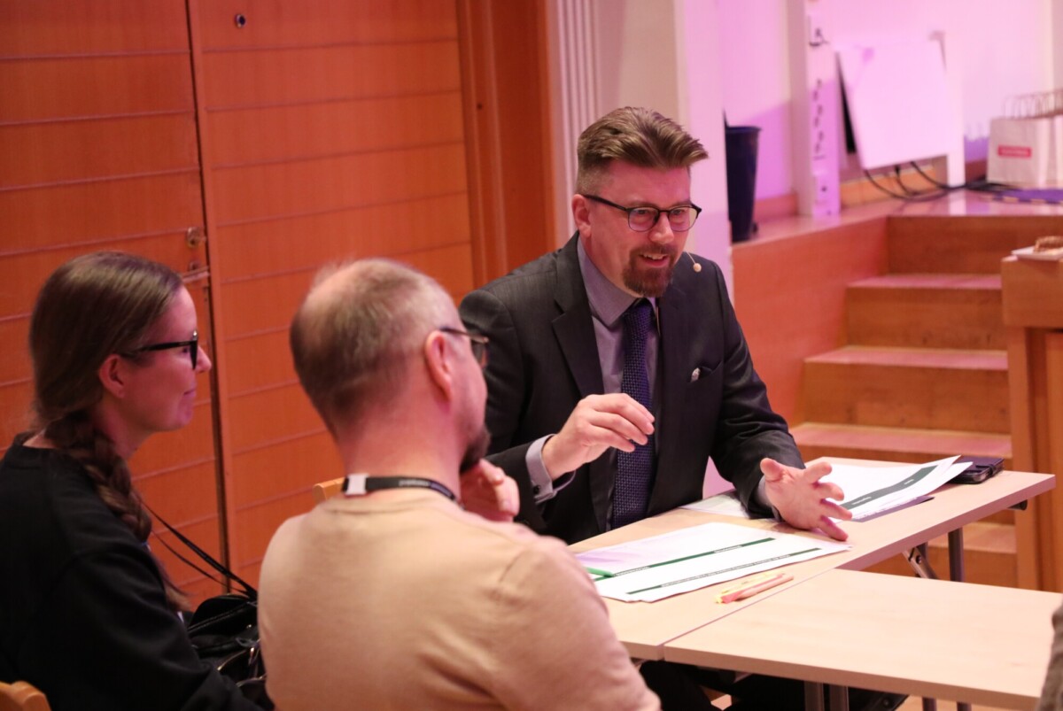 Pukumies ja silmälasipäinen mies puhuu vilkkaasti kahdelle ihmiselle, jotka istuvat häntä vastapäätä pöydässä. Pöydälle on levitetty papereita ja kansioita kirkkaasti valaistussa sisätilassa.