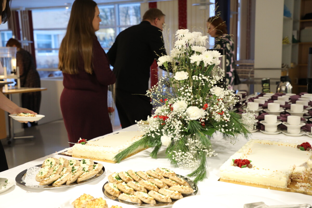 Pöytä, jossa on kakkuja, voileipiä ja suuri kukka-asetelma; taustalla seisoo juhlapukuisia ihmisiä, ja oikealla on pino kuppeja ja lautasia.