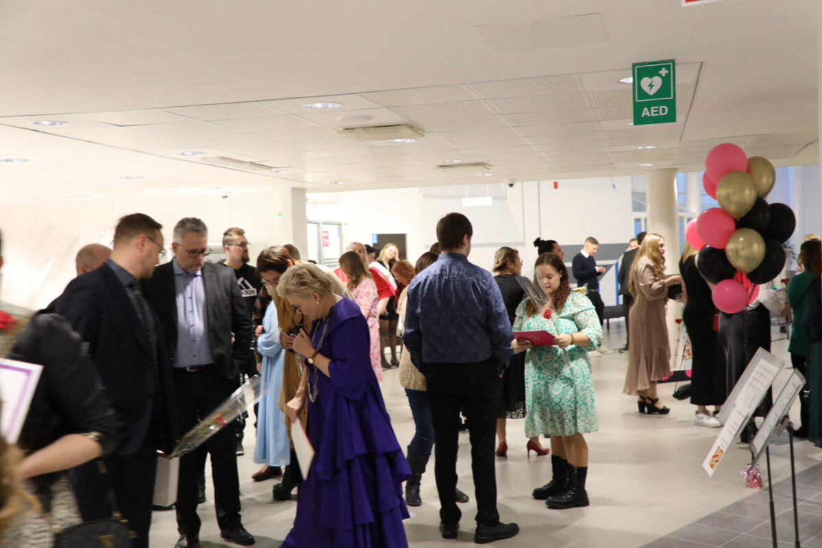 Valoisaan sisätilaan on kokoontunut joukko ihmisiä, joista osa puhuu ja pitää esineitä kädessään. Oikealla näkyy joukko mustia, vaaleanpunaisia ja kultaisia ilmapalloja kyltin vieressä, ja katosta roikkuu AED-kyltti.