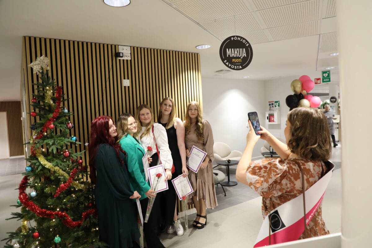 Viisi nuorta naista poseeraa todistusten kanssa koristellun joulukuusen vieressä sisätiloissa, kun toinen nainen, jolla on kaulaliina, ottaa heistä kuvan älypuhelimella. Asetelma vaikuttaa juhlalliselta ja juhlavalta.