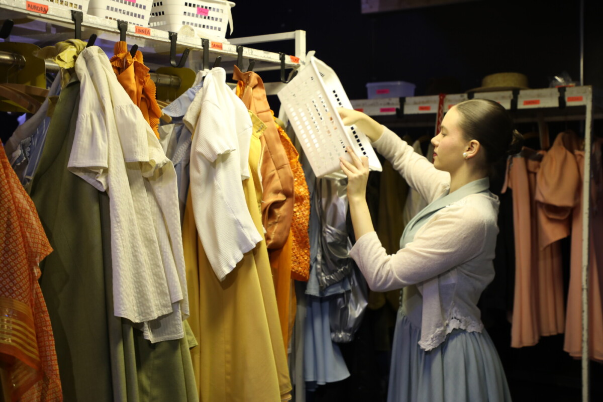 Pukuun pukeutunut nainen seisoo värikkäitä vaatteita täynnä olevan vaatehyllyn edessä järjestelemässä tai hakemassa ylähyllyllä olevaa valkoista muovikoria backstagella tai pukuhuoneessa.