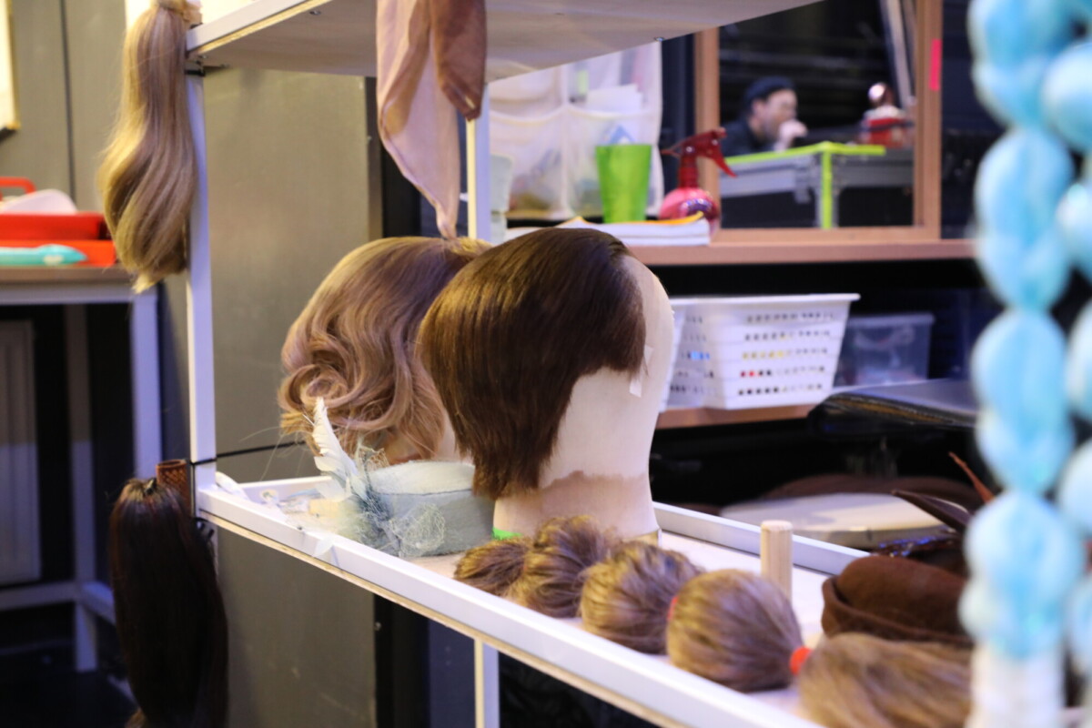 Hyllyllä on kaksi mallinuken päätä, joissa on peruukkeja, hiuskiehkuroita ja hiustenmuotoiluvälineitä. Taustalla on epäselvästi kaksi ihmistä, jotka työskentelevät ilmeisesti backstagella tai pukuhuoneessa.