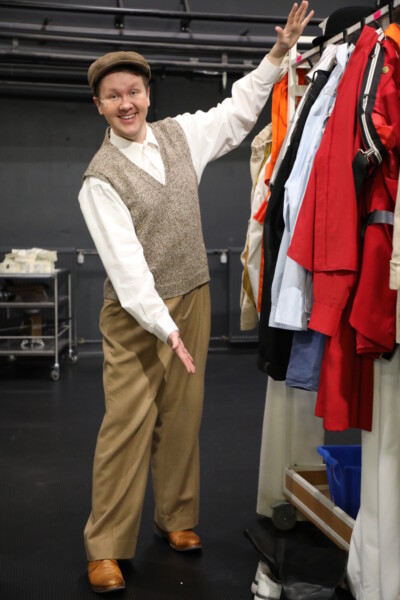 Hymyilevä henkilö, jolla on vintage-tyylinen vaatetus ja litteä lippalakki, osoittaa kohti värikkäiden pukujen hyllyä, joka on ripustettu ripustelineisiin backstagella tai pukuhuoneessa.