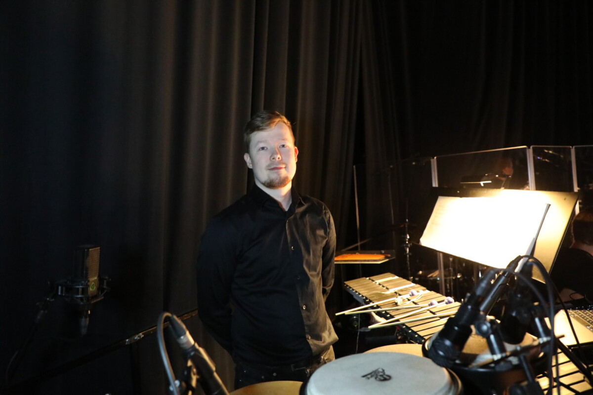 Mustiin vaatteisiin pukeutunut mies seisoo hämärästi valaistulla backstagella lyömäsoittimien ja nuottien vieressä, ja taustalla näkyy mikrofoneja ja musta verho.