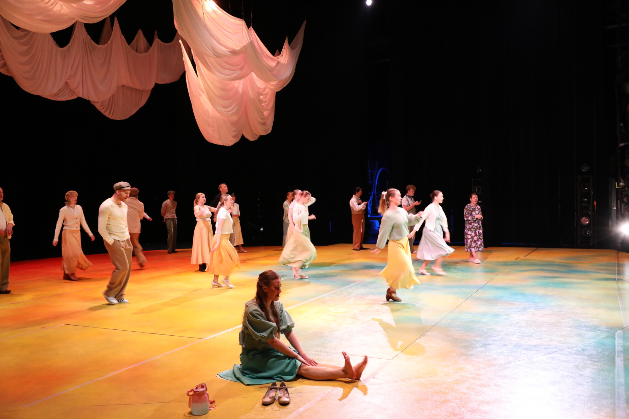 Ryhmä ihmisiä tanssii kirkkaasti valaistulla lavalla, jonka yläpuolella on virtaavia kangaskoristeita, kun taas siniseen mekkoon pukeutunut nainen istuu lattialla etualalla ja säätää kenkiään valonheittimen alla.
