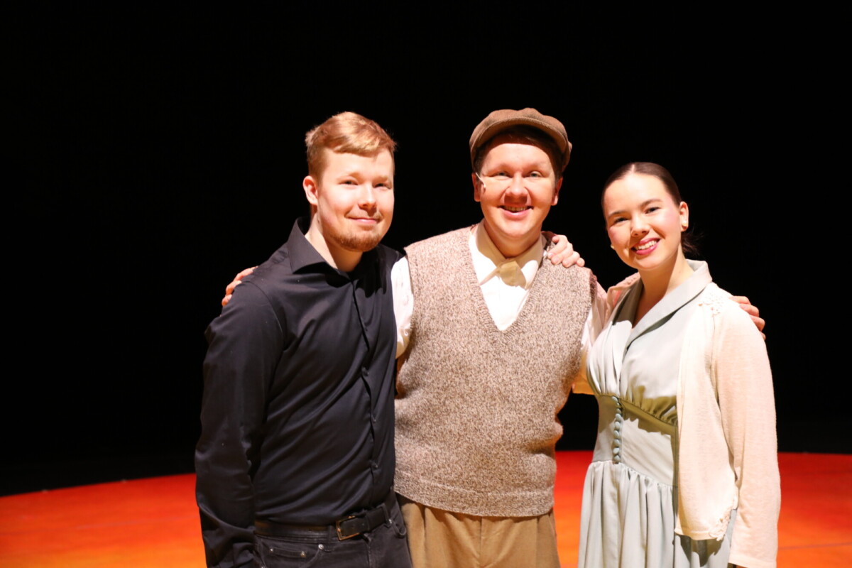 Kolme ihmistä seisoo hymyillen kirkkaasti valaistulla näyttämöllä, jonka tausta on tumma. Keskellä olevalla henkilöllä on litteä lippis ja villaliivi, muilla on musta paita ja vaalea mekko.