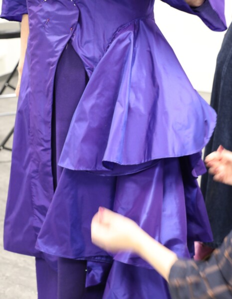 Henkilöllä on yllään kiiltävä, kerroksellinen violetti mekko tai takki, ja toinen henkilö säätää kangasta sumeilla käsillään. Kuvassa keskitytään vaatteen rakenteeseen ja yksityiskohtiin.