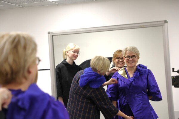 Neljä naista on kirkkaasti valaistussa huoneessa. Yksi nainen säätää toisen violettia mekkoa, kun kaksi muuta katselee ja hymyilee, ja kaikki heijastuvat suuresta peilistä. Ilmapiiri vaikuttaa ystävälliseltä ja yhteistyökykyiseltä.