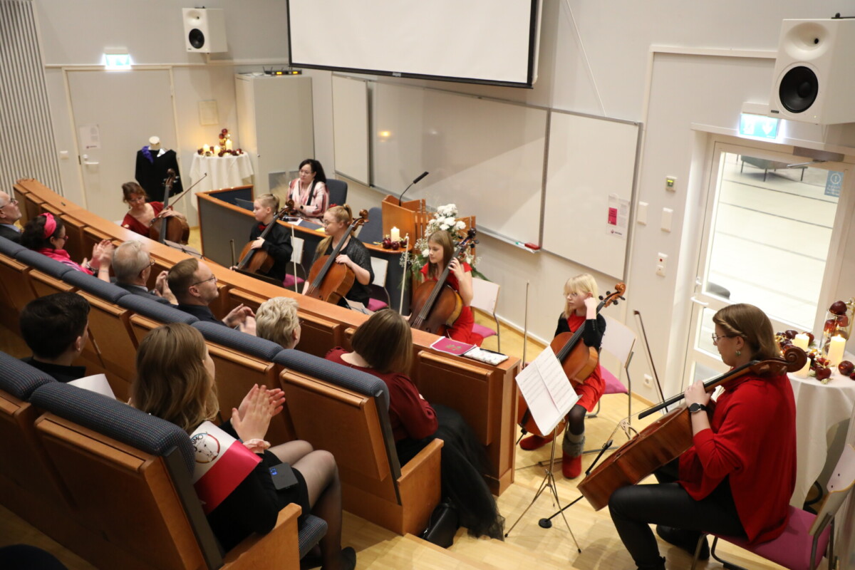 Pieni jousiorkesteri esiintyy luentosalissa yleisölle. Muusikot istuvat sellojen ja nuottitelineiden kanssa, ja läsnäolijat katselevat ja taputtavat. Nainen istuu kirjoituspöydän ääressä valkotaulun ja projektorinäytön edessä.