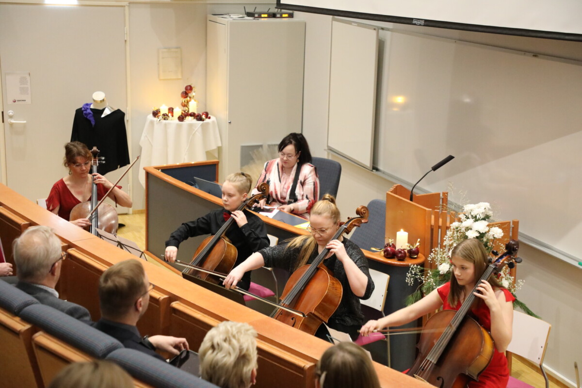 Neljä muusikkoa soittaa jousisoittimia luentosalissa yleisön seuratessa. Nainen istuu taustalla pöydän ääressä taulujen, kynttilöiden ja kukkien ääressä. Tilanne vaikuttaa muodolliselta ja intiimiltä.