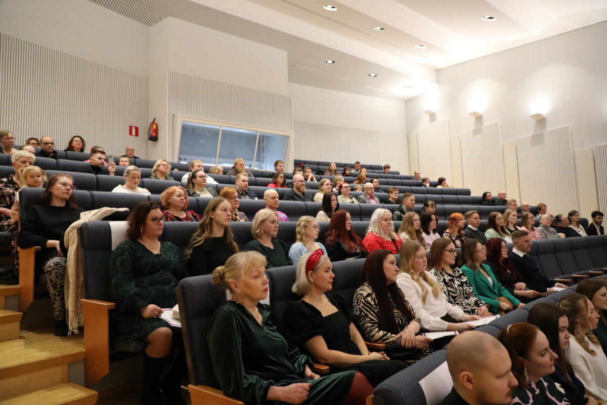 Joukko ihmisiä istuu valoisassa, modernissa luentosalissa tai auditoriossa tarkkaavaisesti porrastetuissa istuinriveissä eteenpäin päin, ikään kuin katselisivat esitystä tai seremoniaa.