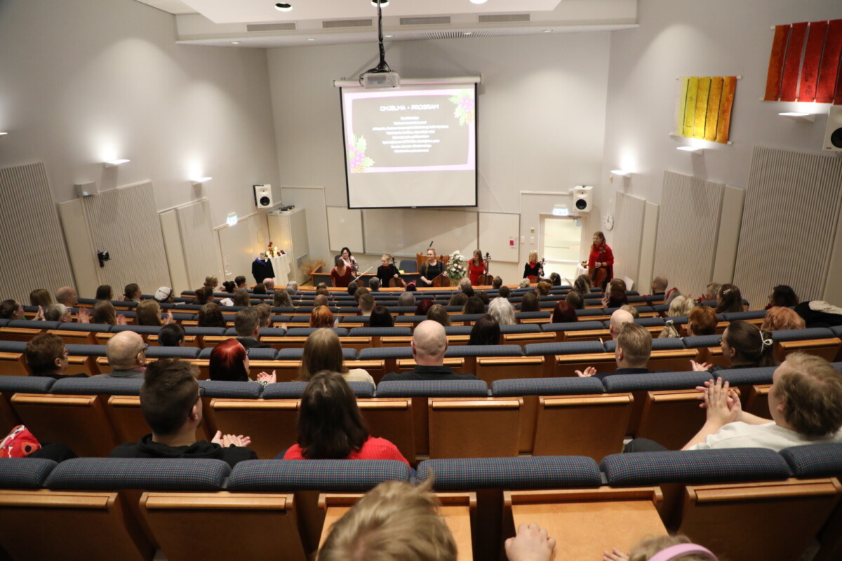 Ryhmä ihmisiä istuu auditoriossa, jossa on lava, jossa useat esittelijät seisovat suuren näytön edessä, jossa näkyy ohjelmaohjelma. Yleisö on tarkkaavainen, jotkut taputtavat.