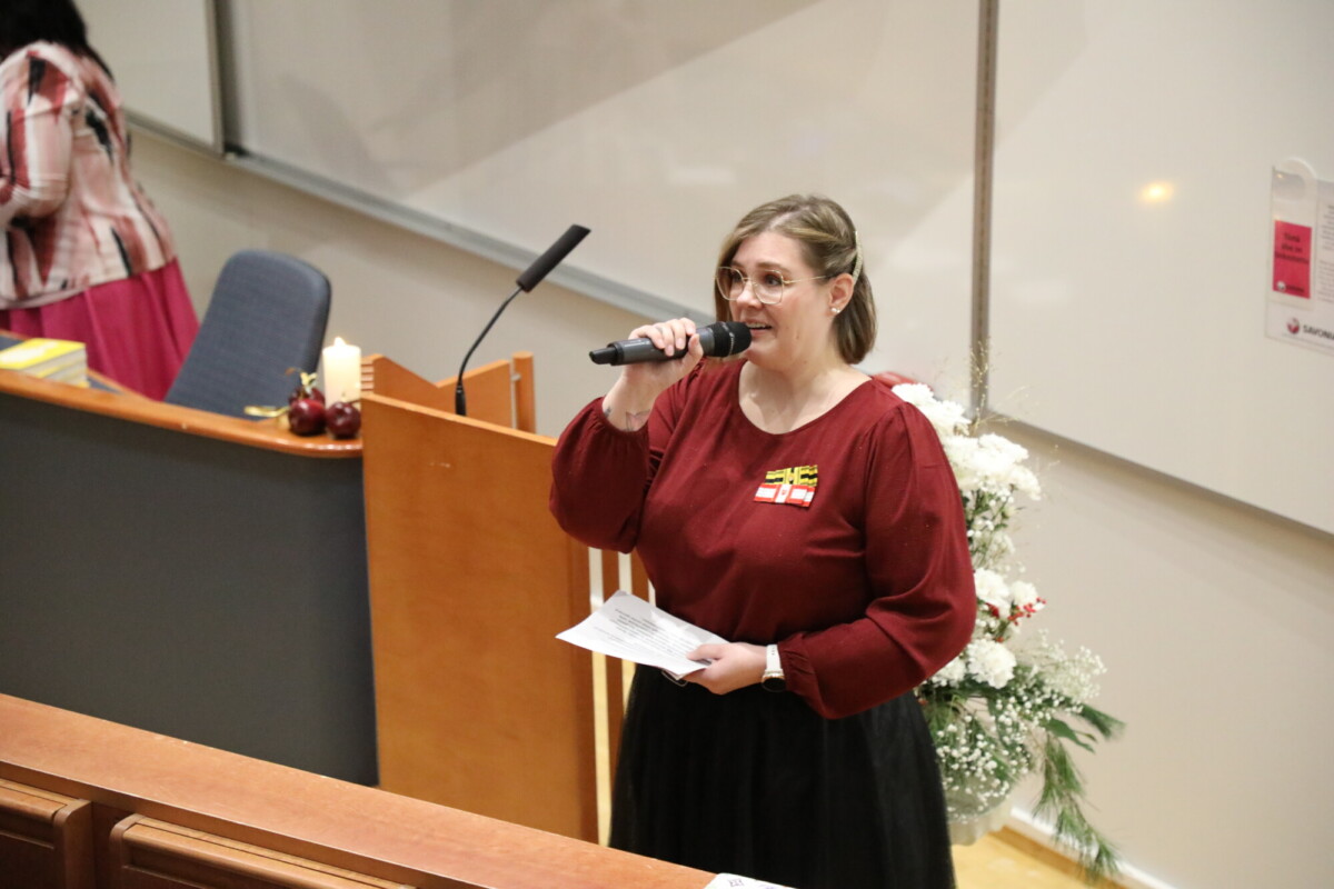 Nainen, jolla on silmälasit ja ruskeankeltainen toppi, puhuu mikrofoniin ja pitää kädessään papereita. Hän seisoo luentosalissa korokkeella. Hänen vieressään olevalla pöydällä näkyy valkoisia kukkia ja omenoita.