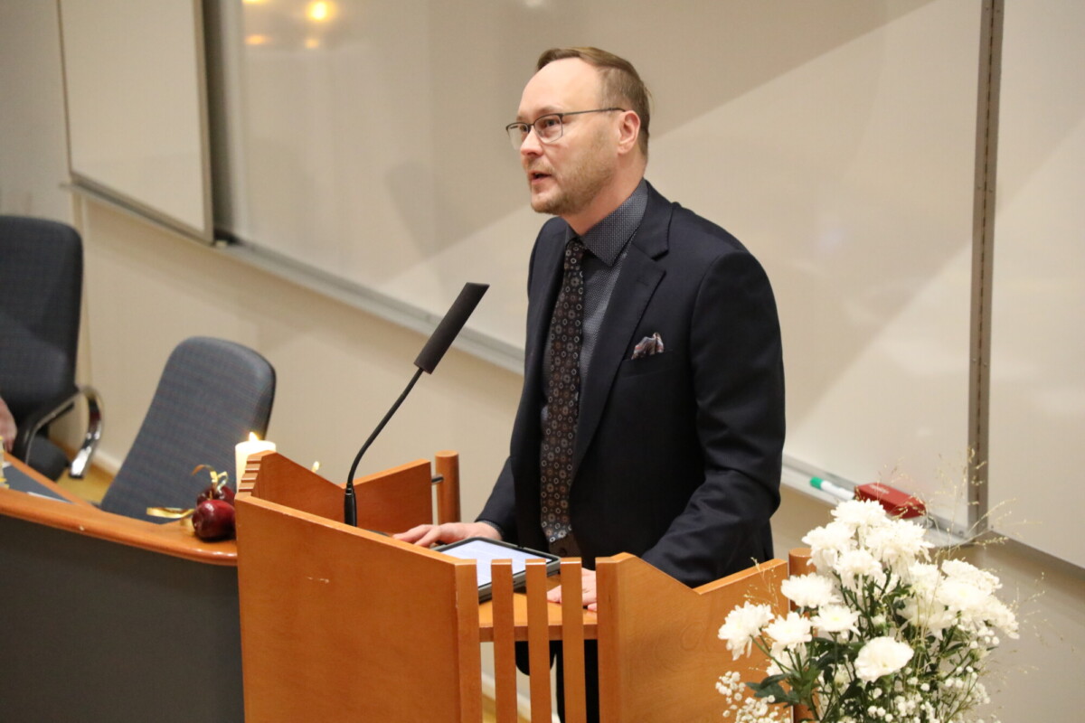 Pukuinen ja silmälasipäinen mies puhuu puisella korokkeella luentosalissa, ja hänen edessään on valkoisia kukkia ja mikrofoni. Ympäristö vaikuttaa muodolliselta, mahdollisesti seremonian tai esityksen aikana.