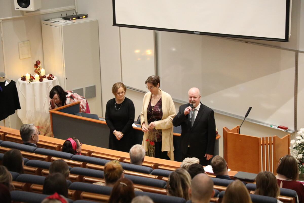 Mies puhuu mikrofoniin luentosalin etuosassa kolmen naisen seurassa. Yleisö istuu, ja taustalla näkyy valkokangas ja koroke.