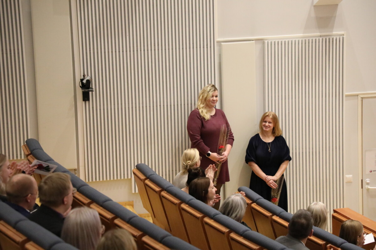 Kaksi naista seisoo ja hymyilee yleisön edessä auditoriossa. Vasemmalla olevalla naisella on kädessään ruusu. Ruskeissa tuolirivissä istuva yleisö näyttää kuuntelevan ja kiinnittävän huomiota.