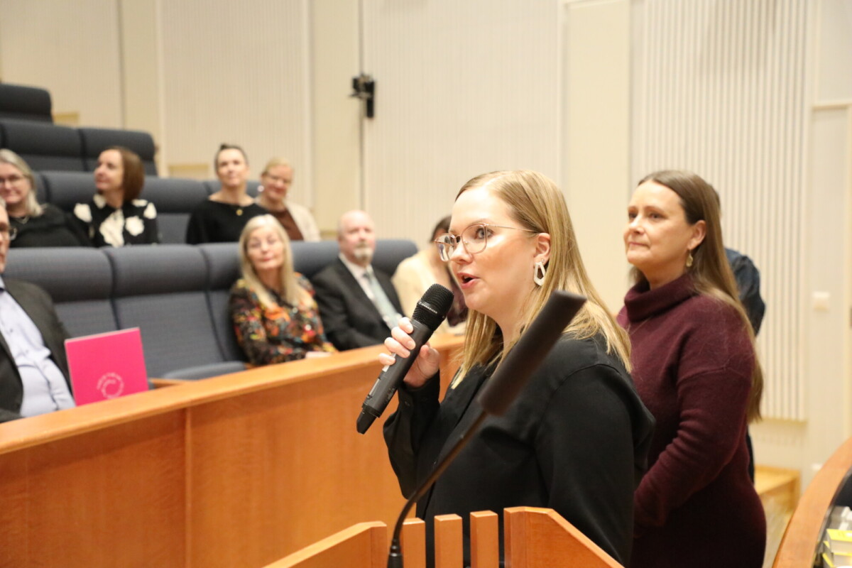 Mikrofonia pitelevä nainen puhuu luentosalissa, ja hänen vieressään seisoo toinen nainen ja taustalla istuu useita ihmisiä, jotka katselevat ja kuuntelevat tarkkaavaisesti.
