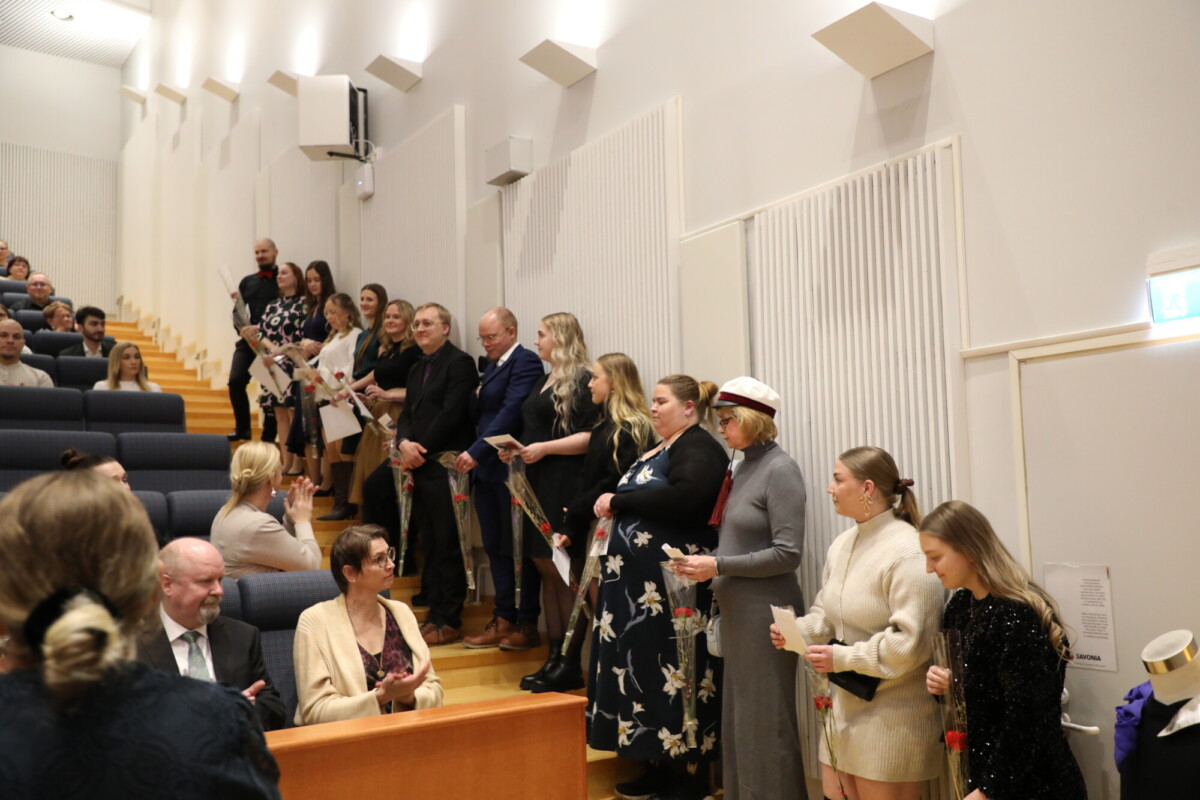 Joukko ihmisiä seisoo rivissä luentosalin seinää pitkin, kädessään papereita ja kukkia, kun taas muut istuvat riveissä ja katselevat tapahtumia. Sali on kirkkaasti valaistu ja siinä on porrastetut istuimet.