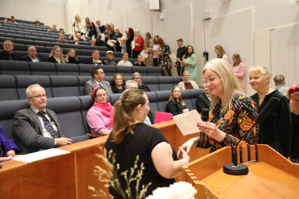 Nainen ojentaa todistuksen toiselle naiselle luentosalin korokkeella. Ihmiset istuvat ja seisovat, monet hymyilevät ja katselevat vaihtoa. Tunnelma viittaa viralliseen tai juhlalliseen tapahtumaan.