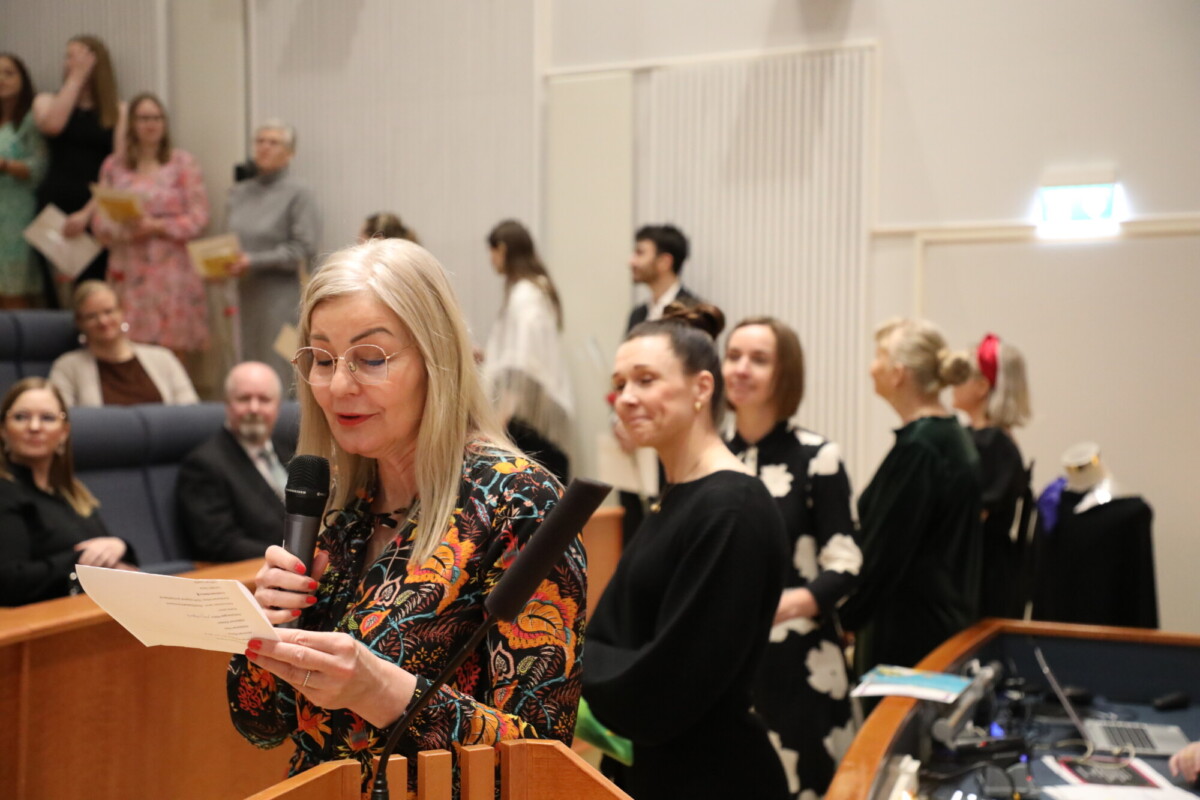 Pitkät vaaleat hiukset ja silmälasit päässä oleva nainen lukee paperia mikrofoniin luentosalin korokkeella, ja taustalla seisoo ja istuu ihmisiä, joilla on kädessään papereita ja jotka hymyilevät.