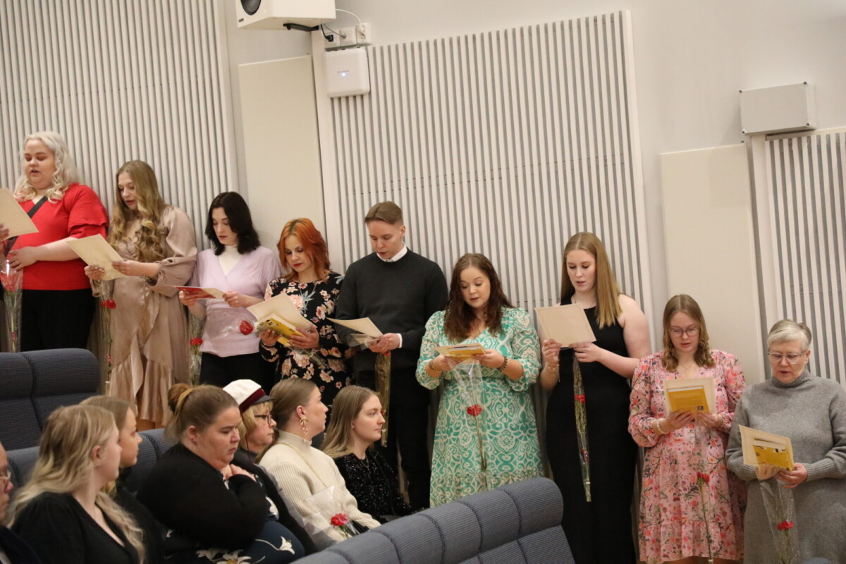 Joukko ihmisiä seisoo rivissä, kädessään papereita ja ruusuja, laulaa tai lukee. Jotkut istuvat etualalla ja kuuntelevat tarkkaavaisesti. Ympäristö näyttää olevan sisätiloissa järjestettävä tapahtuma tai seremonia, jossa on raidalliset seinäpaneelit.