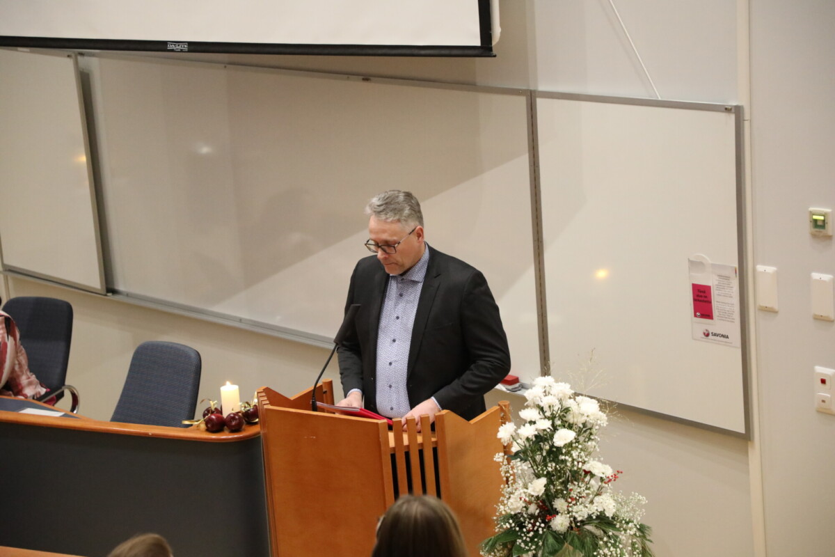 Pukumies seisoo puisen korokkeen ääressä ja lukee luentosalissa esitelmää. Valkoiset kukat ja palavat kynttilät koristavat koroketta, ja ihmiset istuvat kuuntelemassa häntä. Valkoiset taulut peittävät seinät hänen takanaan.