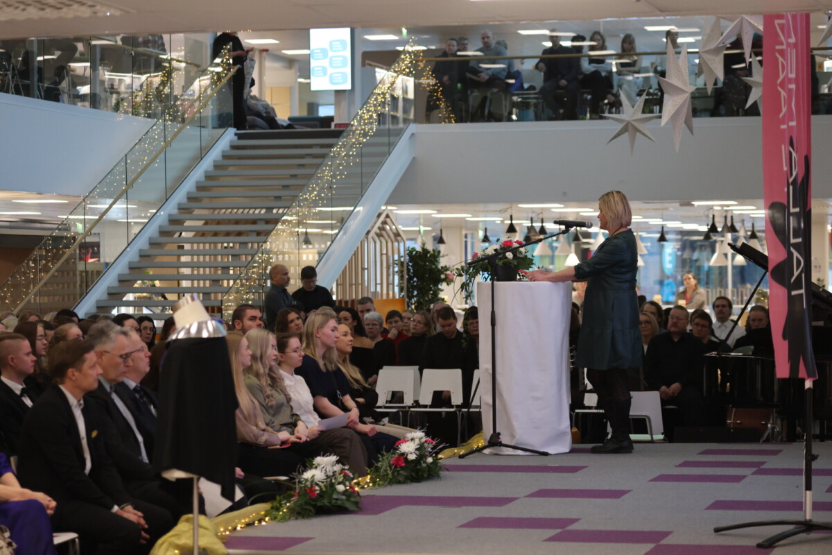 Nainen seisoo korokkeella puhumassa istuvalle yleisölle modernissa, sisustetussa sisätiloissa, joiden taustalla on lomavaloja, kukkia ja suuret portaat. Ihmiset katselevat molemmista kerroksista.