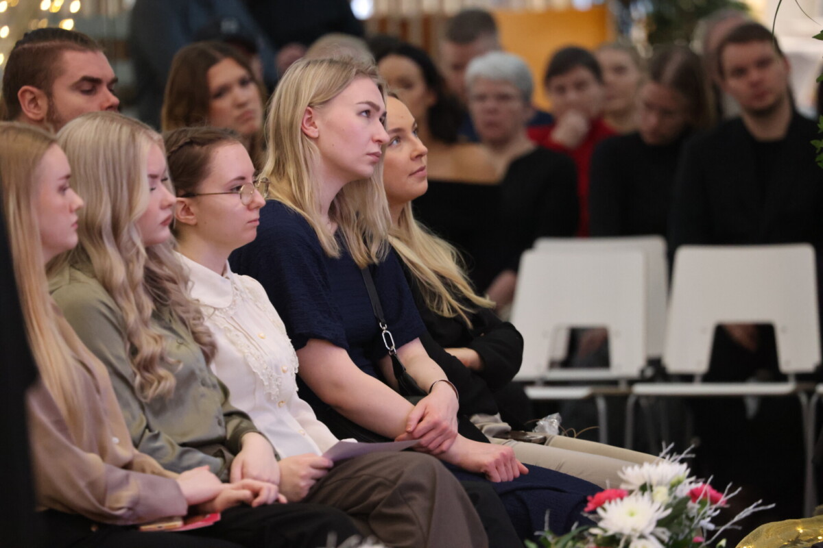 Ryhmä ihmisiä istuu tiiviisti rivissä sisätiloissa järjestettävässä tapahtumassa ja kuuntelee keskittyneesti. Jotkut pitävät papereita sylissään. Etualalla on kukkia, ja taustalla on lisää osallistujia.