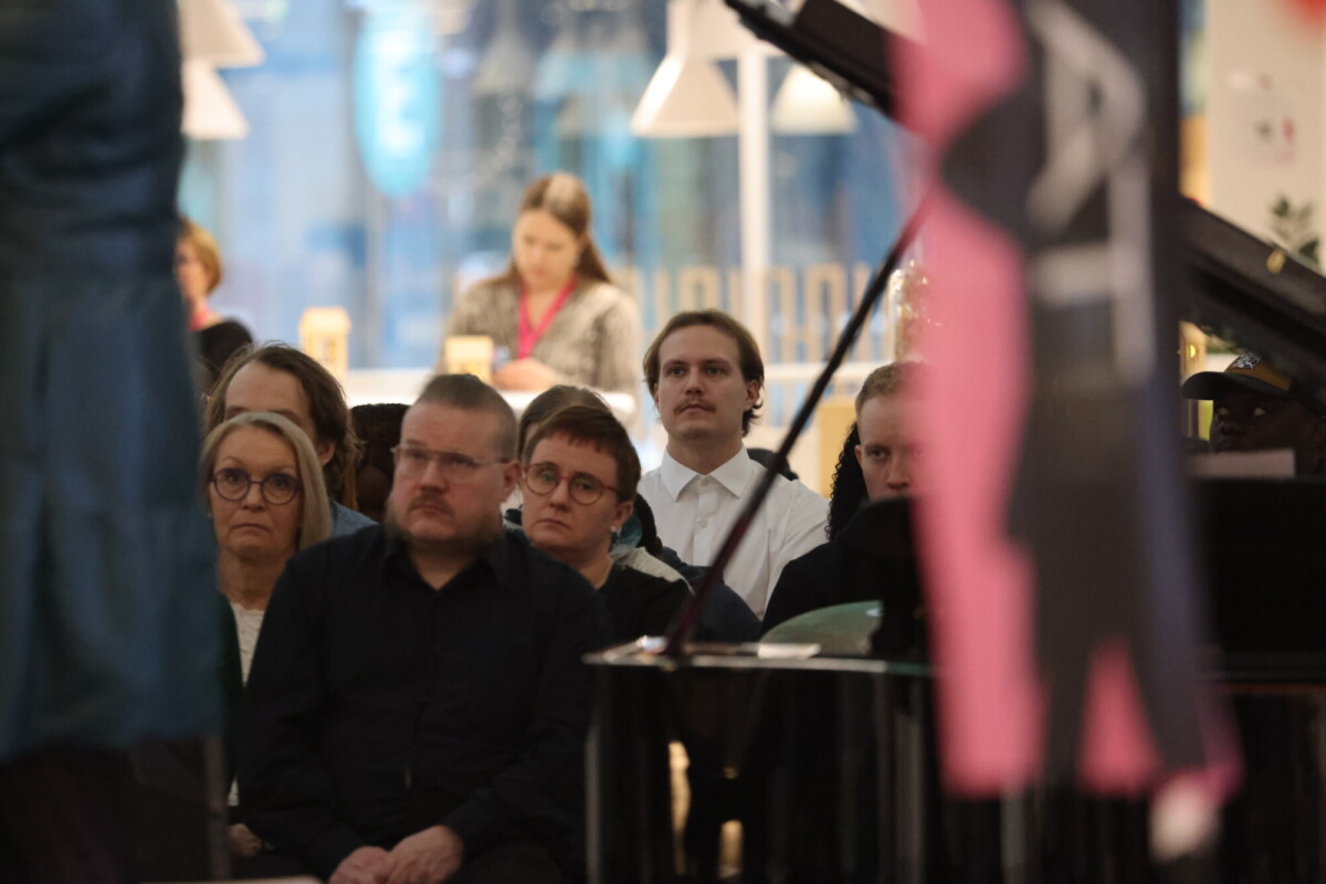 Ryhmä ihmisiä istuu sisätiloissa, tarkkaavaisesti eteenpäin katsoen. Taustalla nainen seisoo tiskin ääressä työskentelemässä. Etualalla on epäselvä osa pianosta ja osittain näkyvä lippu.