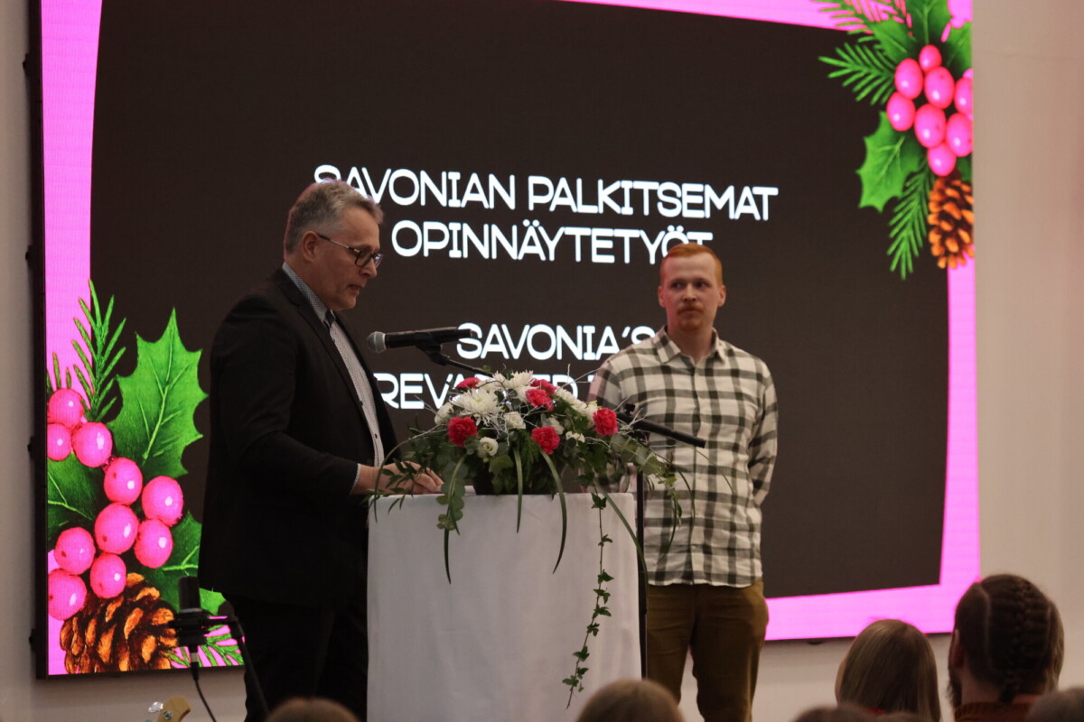 Kaksi miestä seisoo lavalla, jossa on kukkien koristama koroke. Heidän takanaan on suuri valkokangas, jossa näkyy suomenkielistä tekstiä ja juhlallinen reunus, jossa on joulutähtiä, marjoja ja käpyjä. Etualalla näkyy yleisöä.