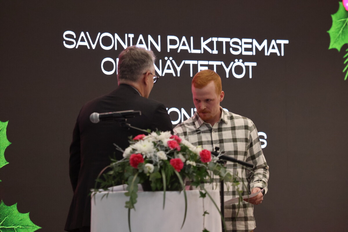 Kaksi miestä seisoo kukkakorokkeen edessä vastakkain. Heidän takanaan näkyy tummalla taustalla suuri suomenkielinen teksti. Tunnelma vaikuttaa muodolliselta, mikä viittaa palkitsemis- tai tunnustustilaisuuteen.