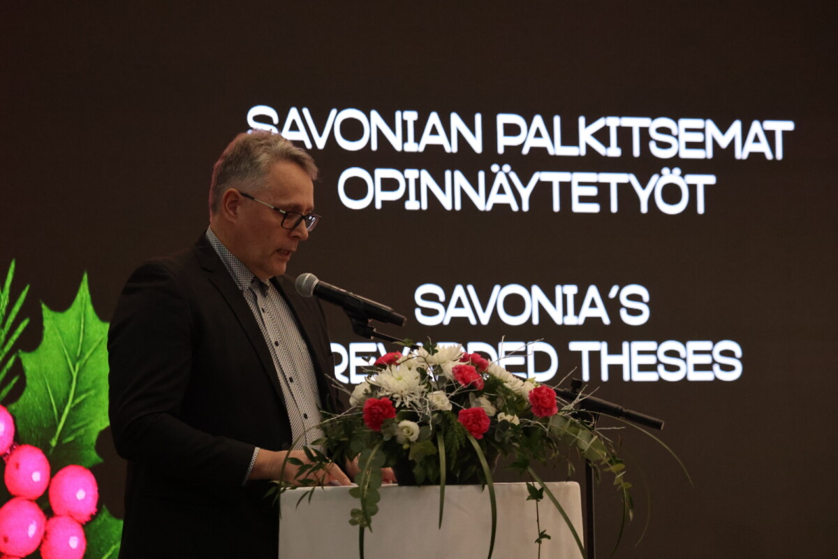 Silmälasipäinen ja pukumies puhuu kukkien koristelemalla korokkeella, hänen takanaan näytöllä teksti SAVONIAN PALKITSEMAT OPINNÄYTETYÖT SAVONIAS PALKITSEE TESTA.