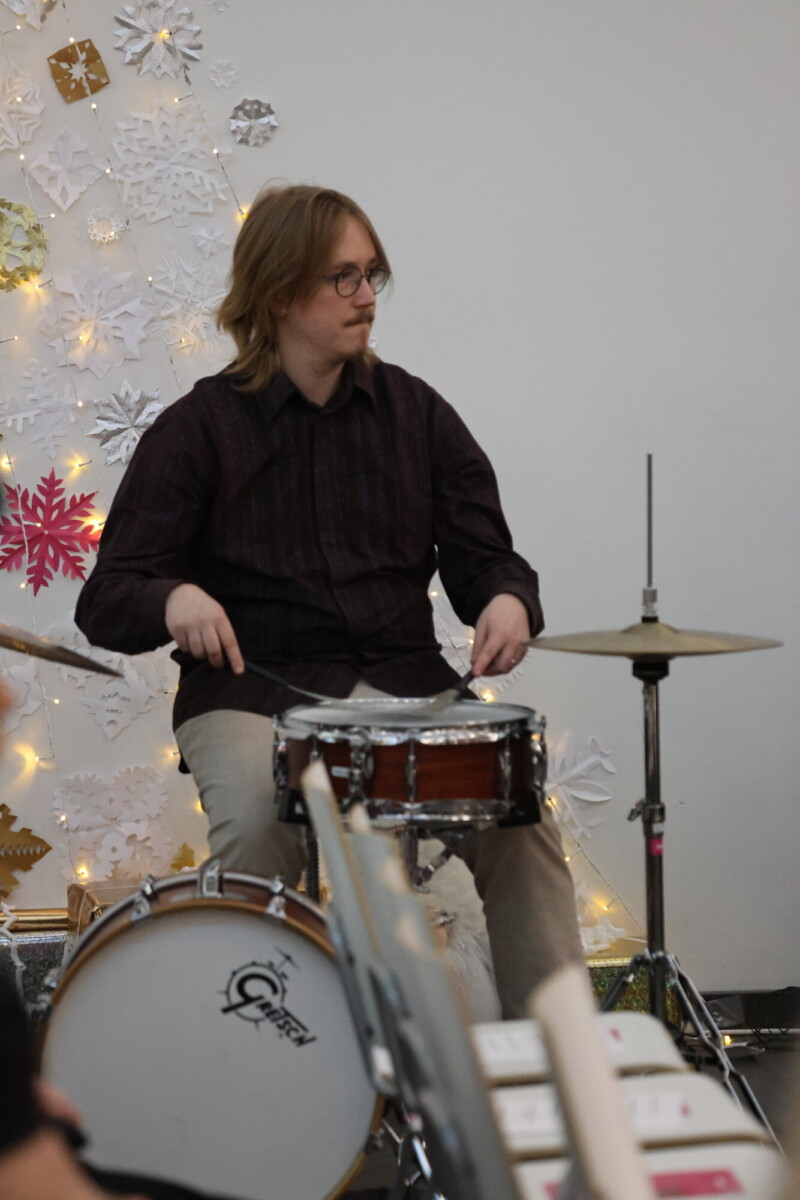 Olkapäiden pituiset hiukset ja silmälasit päässä oleva henkilö soittaa rumpuja paperilumihiutaleilla ja valonjohdoilla koristellun seinän edessä.