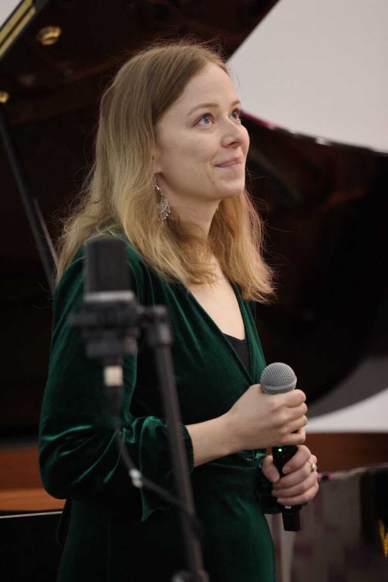 Pitkät vaaleat hiukset omaava nainen, jolla on vihreä samettimekko ja mikrofoni kädessään, seisoo pianon ja mikrofonitelineen edessä katsoen hieman ylöspäin mietteliäänä.