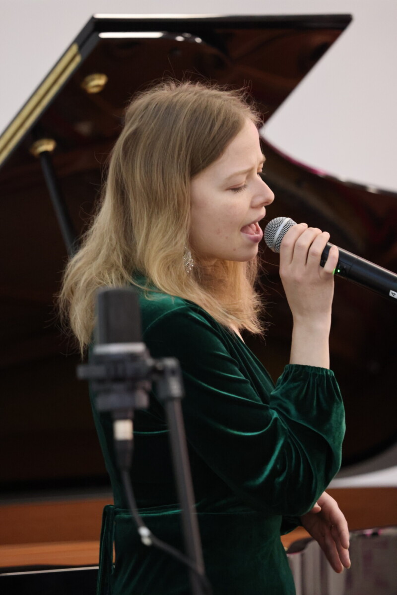 Vihreään samettimekkoon pukeutunut nuori nainen laulaa mikrofoniin, taustalla on flyygeli ja etualalla mikrofoniteline.