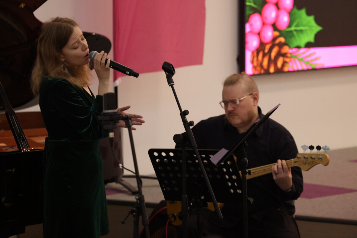 Vihreään mekkoon pukeutunut nainen laulaa mikrofoniin, kun hänen vieressään oleva mies soittaa sähkökitaraa. Heidän takanaan olevalla ruudulla näkyy juhlava tausta, jossa on joulutähtiä ja käpyjä.