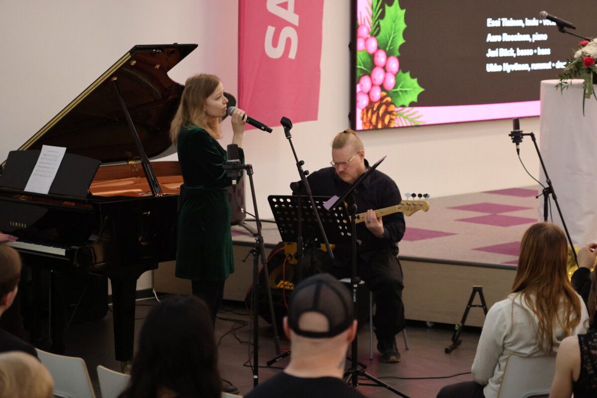 Nainen laulaa mikrofoniin ja mies soittaa kitaraa hänen vieressään; lähellä on piano, ja yleisö seuraa heidän esitystään. Taustalla olevalla ruudulla näkyy juhlallinen diakuva, jossa on joulutähtiä.