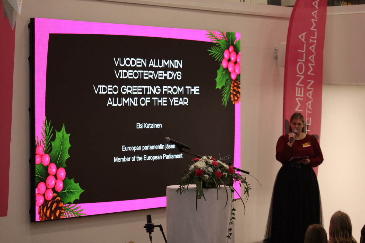 Nainen seisoo korokkeella, jossa on kukkia, ja puhuu näytön edessä, jossa näkyy vuoden alumnitervehdys suomeksi ja englanniksi, ja näytöllä on juhlallisia käpyornamentteja.